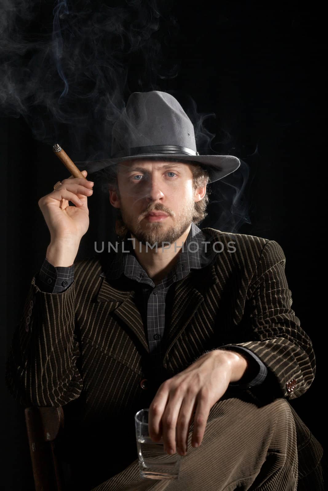 A  man in a grey hat sitting with a glass of alcohol and cigar