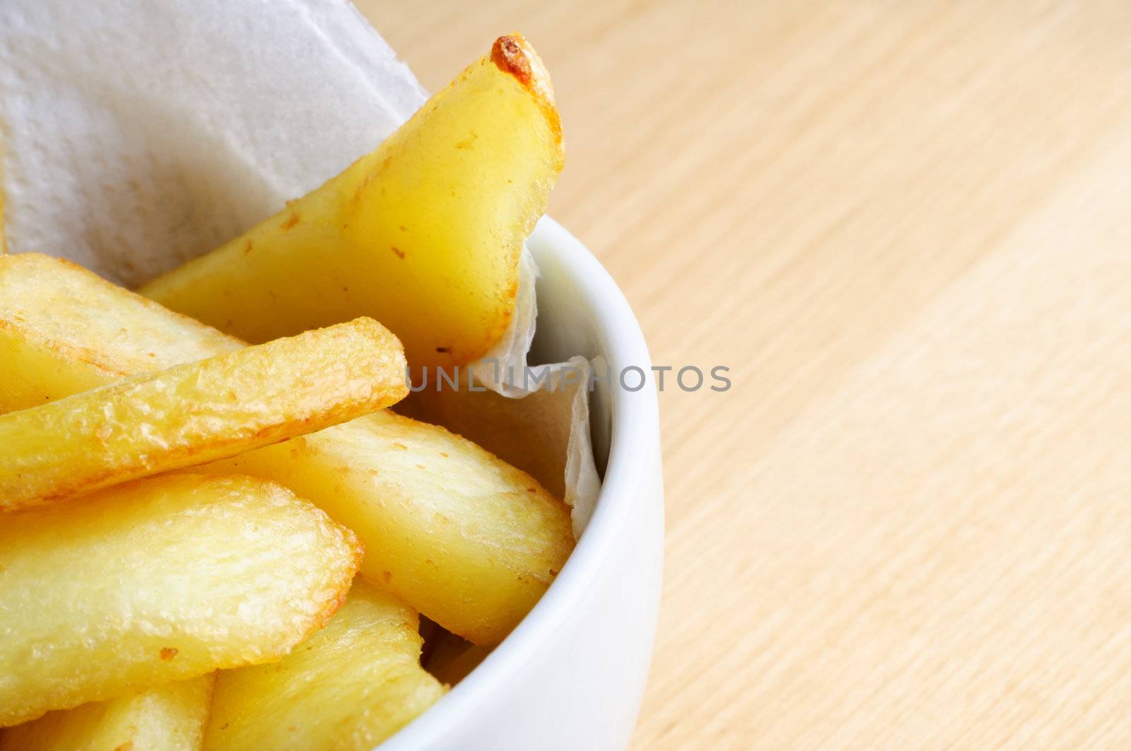 Close up (macro) of a bowl of chips (french fries) nestling on absorbent paper in a white bowl.  Wooden table surface provides copy space to the right.