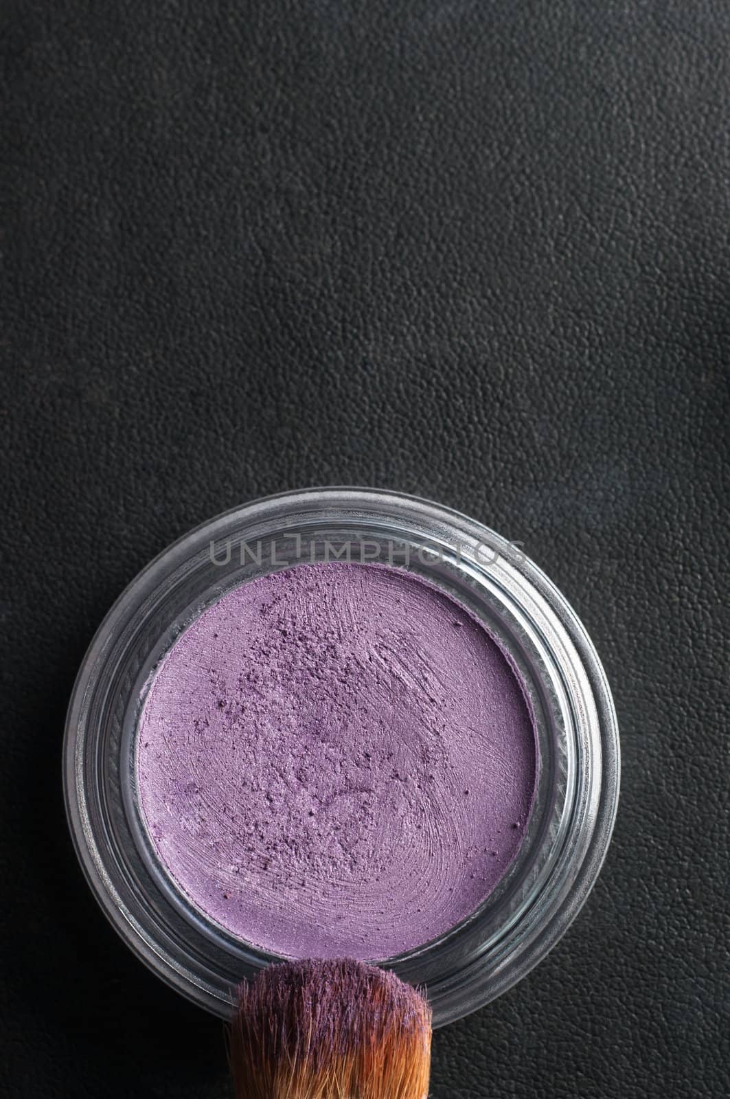 Overhead close up (macro) of an opened pot of purple eye shadow, with make up brush coated in powder on black leatherette background.