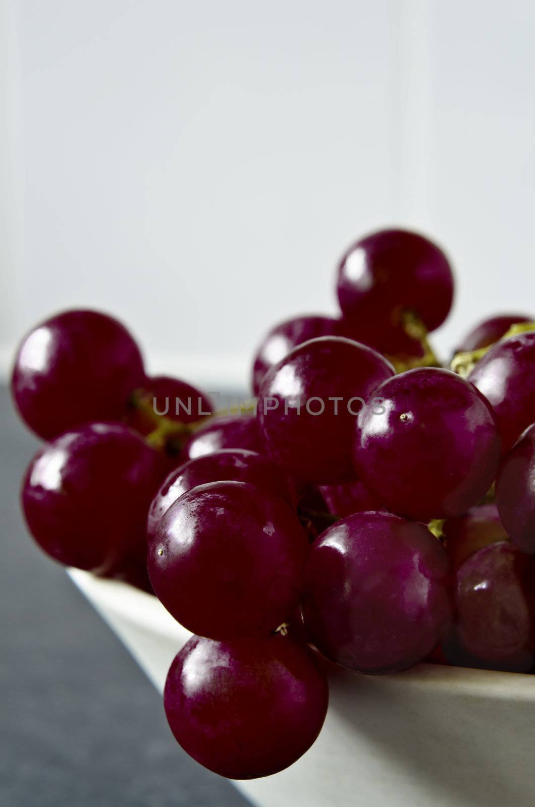 Close up of shiny, wet purple grapes, tumbling over the edge of a bowl on grey granite kitchen surface with white kitchen tiles in soft focus background.  Not their original colour, re-tinted in post-production.