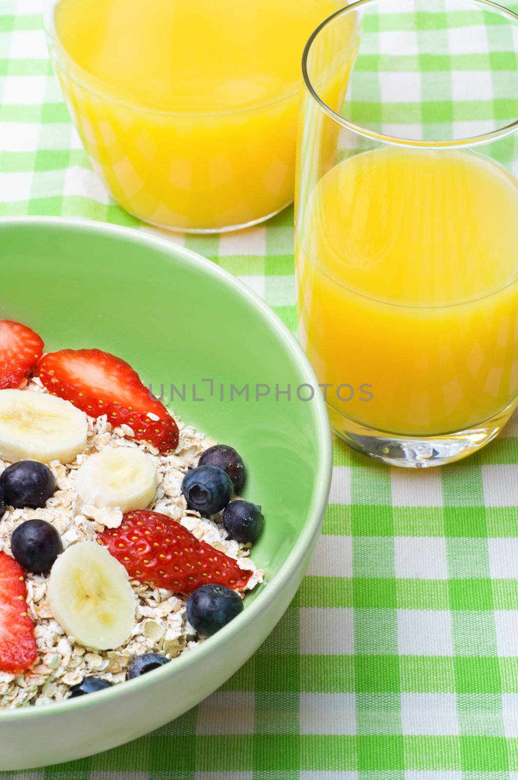 A breakfast setting with a bowl of fresh fruit and oats muesli and orange juice on a green and white gingham tablecloth.