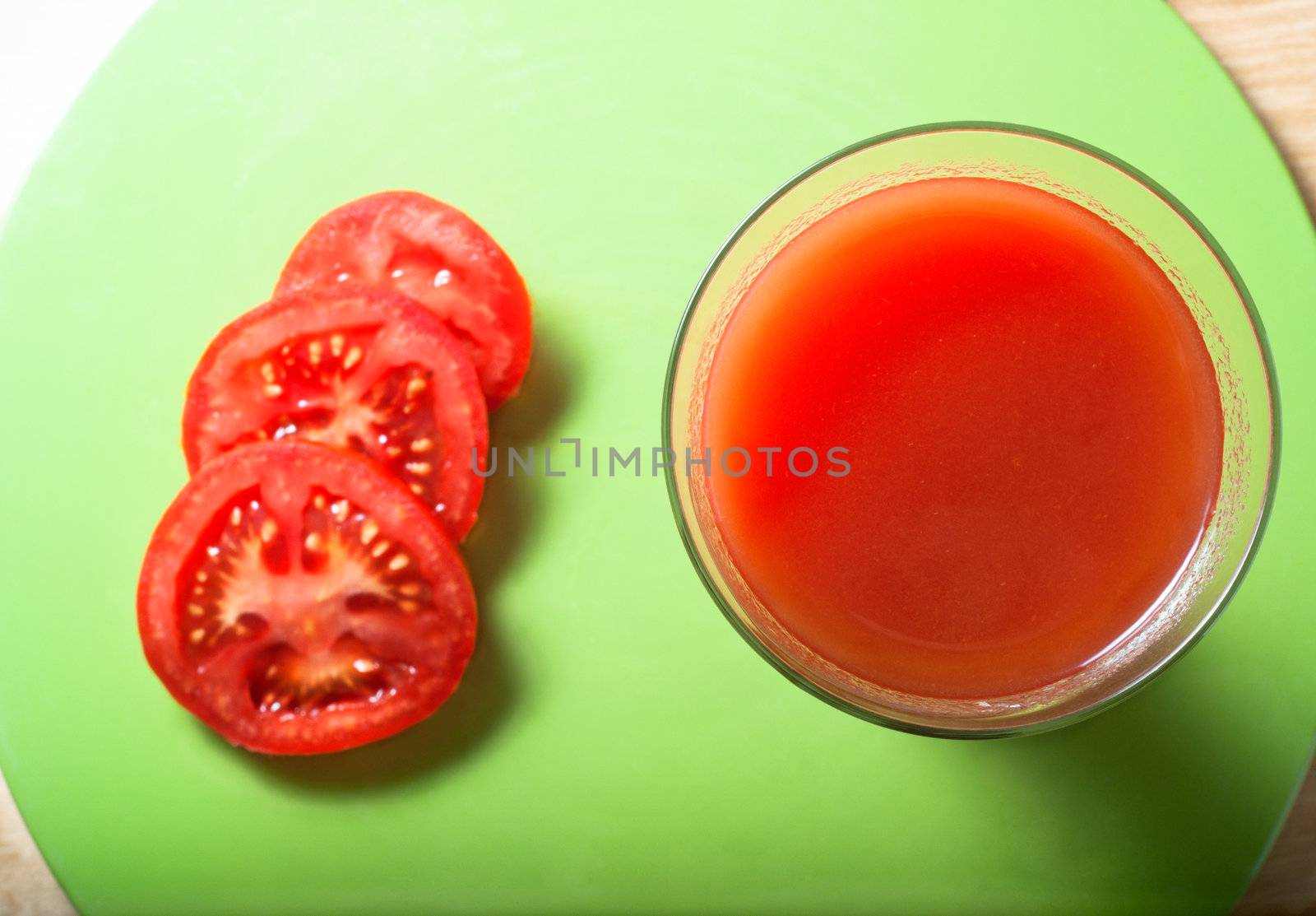 Overhead shot of tomato juice in a long, tall glass beside three slices on a green circular mat, resting on   wood table.  