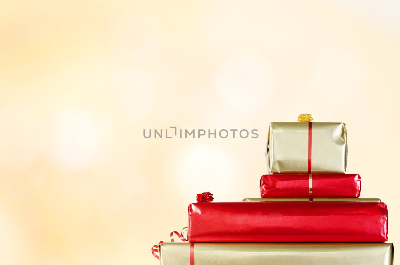 A stack of Christmas parcels, wrapped in gold and red paper and decorated with ribbon and rosettes, against a yellow toned bokeh background.