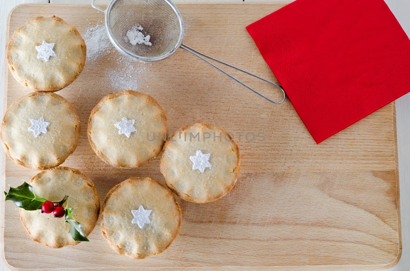 Overhead shot of mince pies decorated with holly sprig and sifted icing sugar in star shapes.  An old tea straining sieve sits on wooden board with sprinkled icing sugar. Red napkin on upper right.