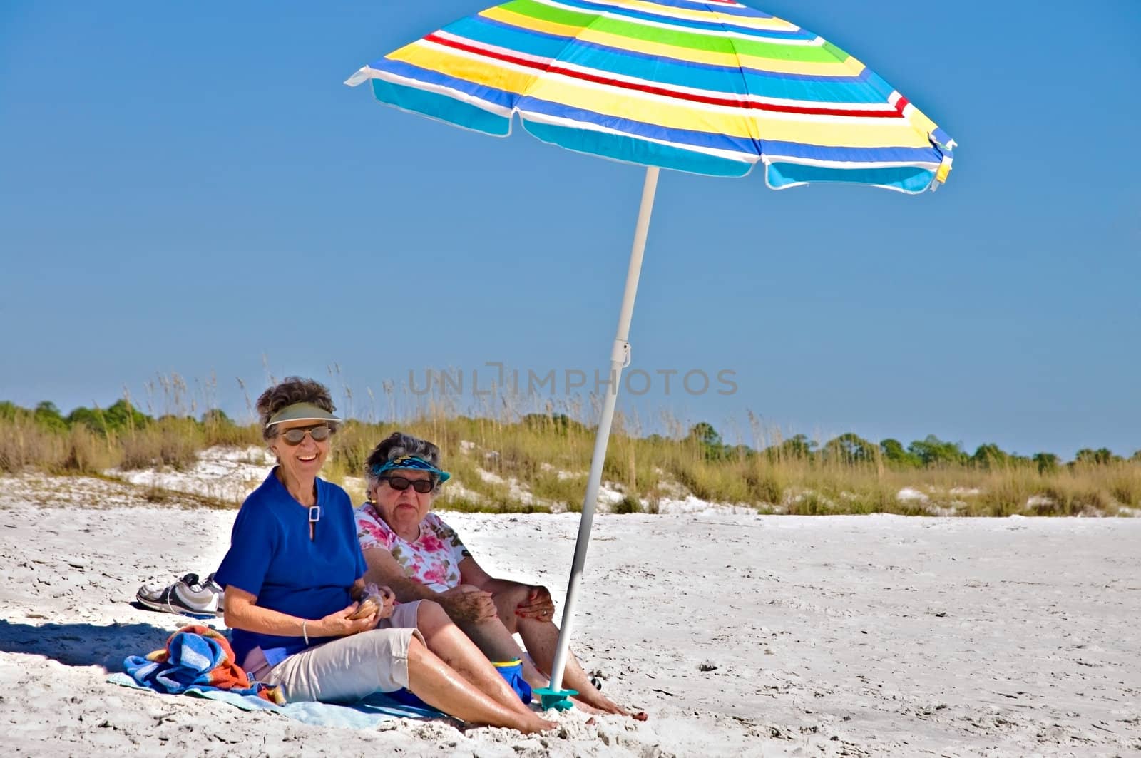 Two elderly women sitting under an umbrella at the beach.