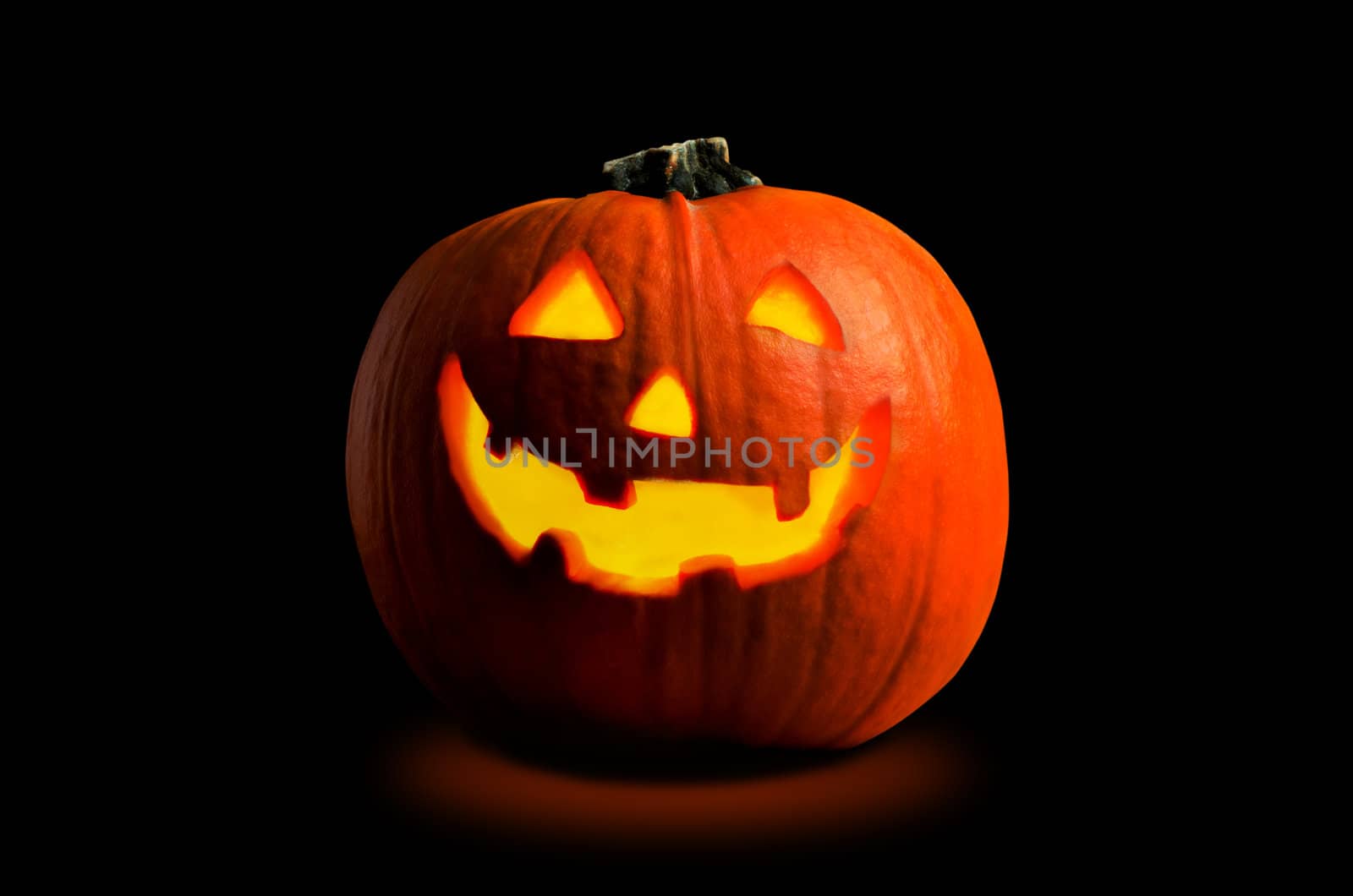 Photograph of a Halloween pumpkin (Jack O' Lantern), with carved face, lit by candlight from the inside on a black background with orange glow beneath.