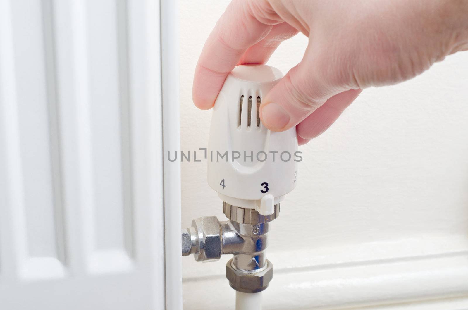 Close up of a hand turning a radiator knob either up or down.  Steel fittings and part of radiator visible, with cream painted wall and skirting board in the background.