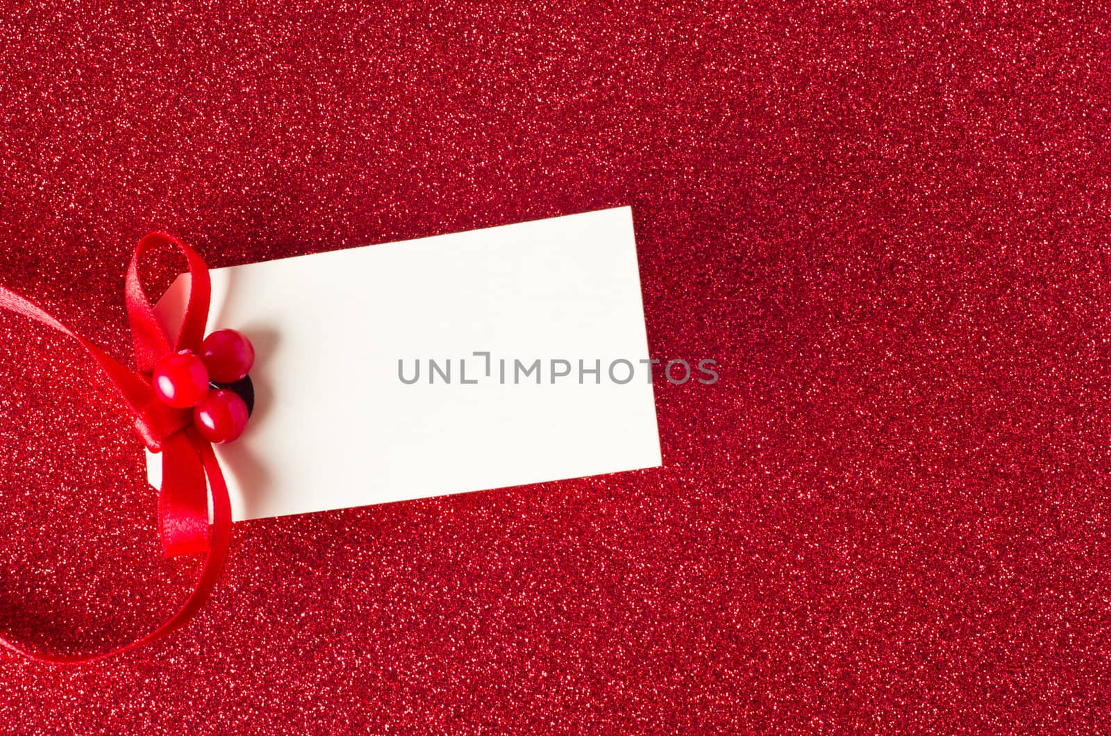 A blank Christmas gift tag, decorated with red ribbon and artifical holly berries, on a sparkly red glitter background.  Copy space on tag.