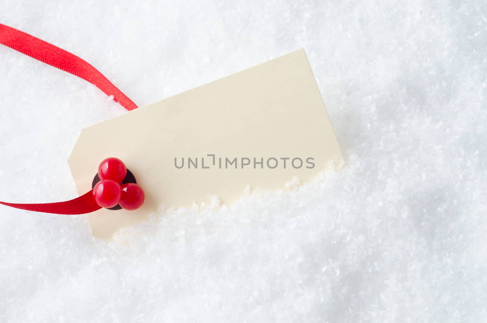 A blank Christmas gift tag, attached to a red ribbon and decorated with plastic red holly berries, nestling in artificial snow.  Copy space on cream coloured tag.