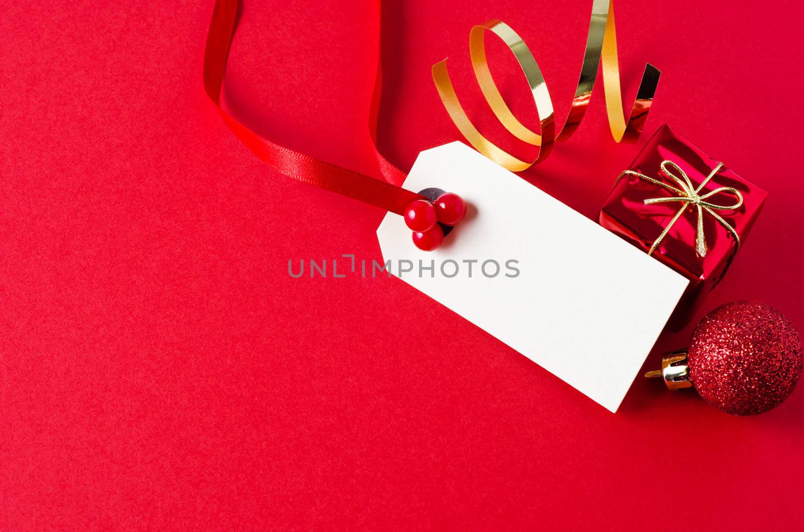 A blank Christmas gift tag, decorated with red ribbon and artifical holly berries, on a matte red background with shiny small gift box, glittery bauble, and gold foil spiral.  Copy space on tag and to it's left.