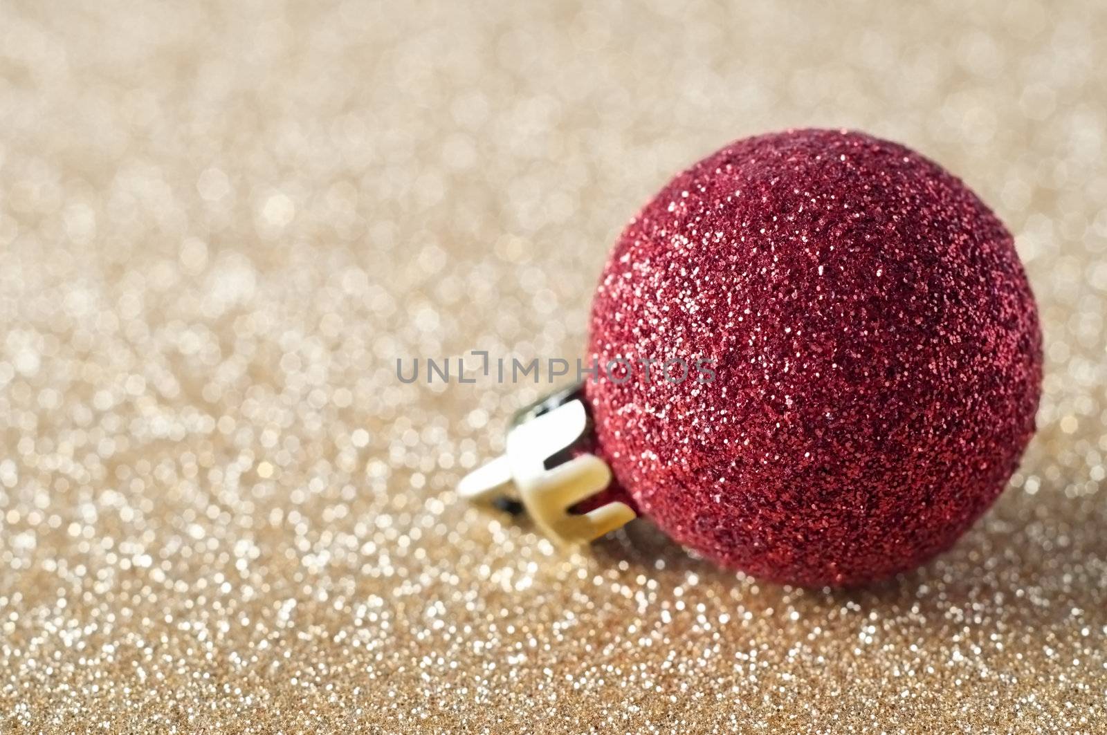 A single, sparkly red bauble, coated in glitter, resting on a gold glitter background that softens into soft focus bokeh in the background.  Copy space to left.