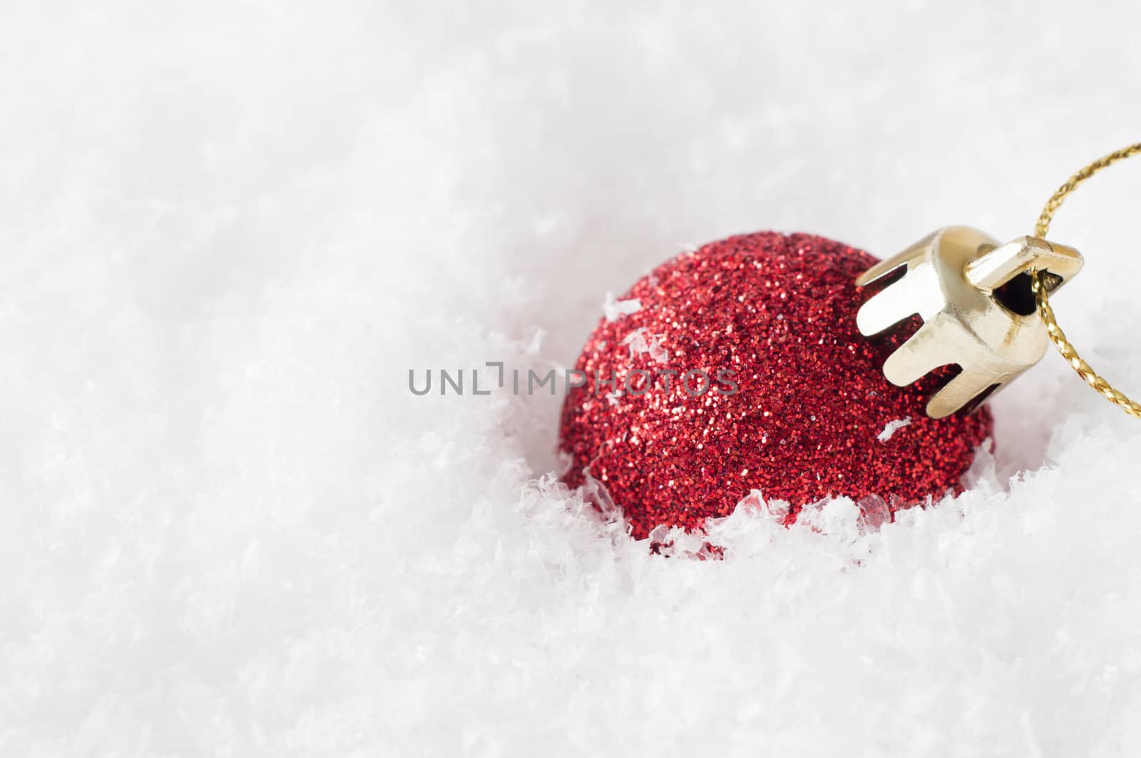 A solitary shiny red Christmas bauble, coated in glitter with gold clasp and string, sunk into artificial white snow and leaning to the right.  Copy space to left.