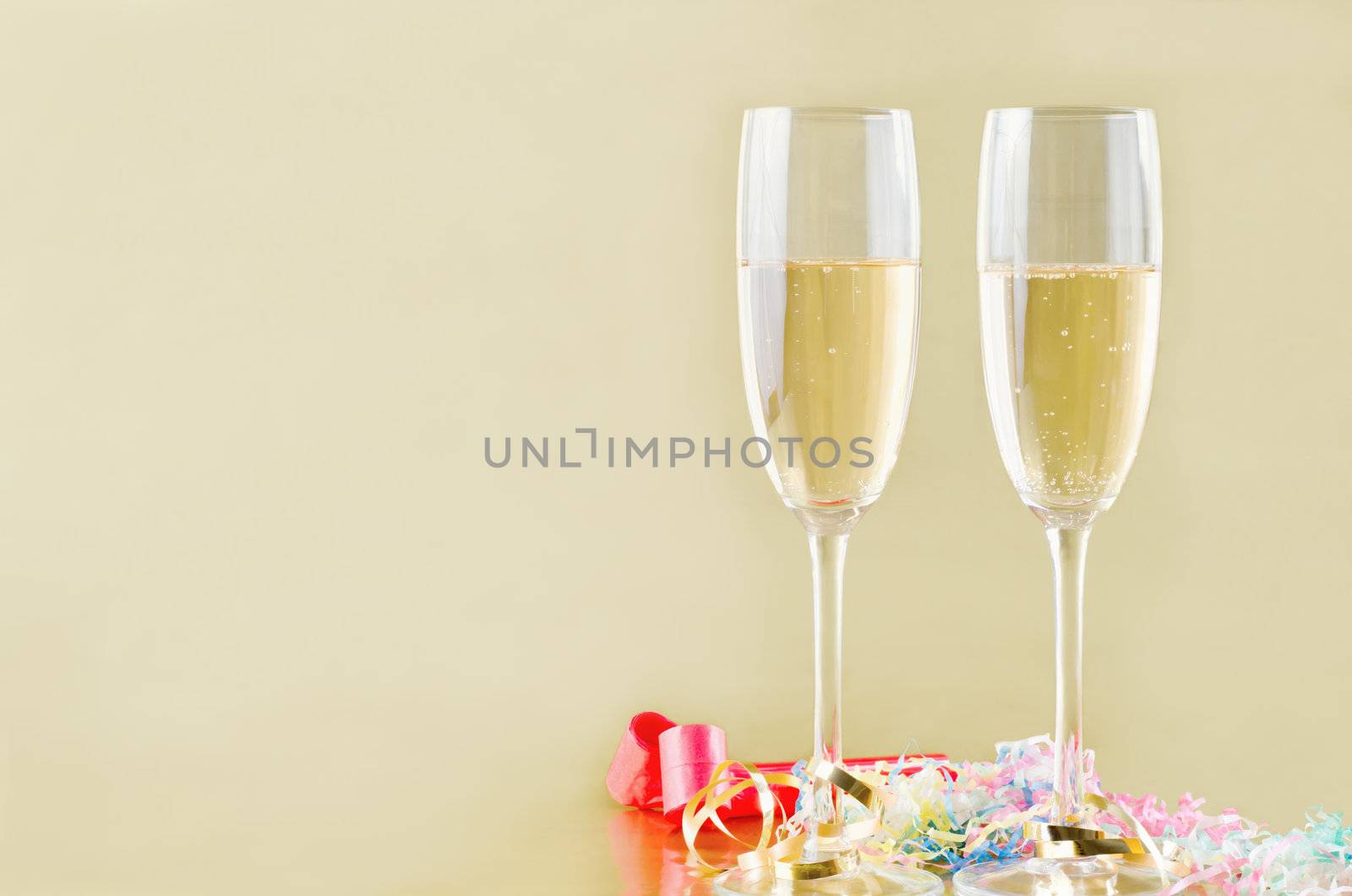 Two fluted champagne glasses with bubbles rising on a gold background with popper streamers and party horn blowers on the surface below.  Copy space to the left.