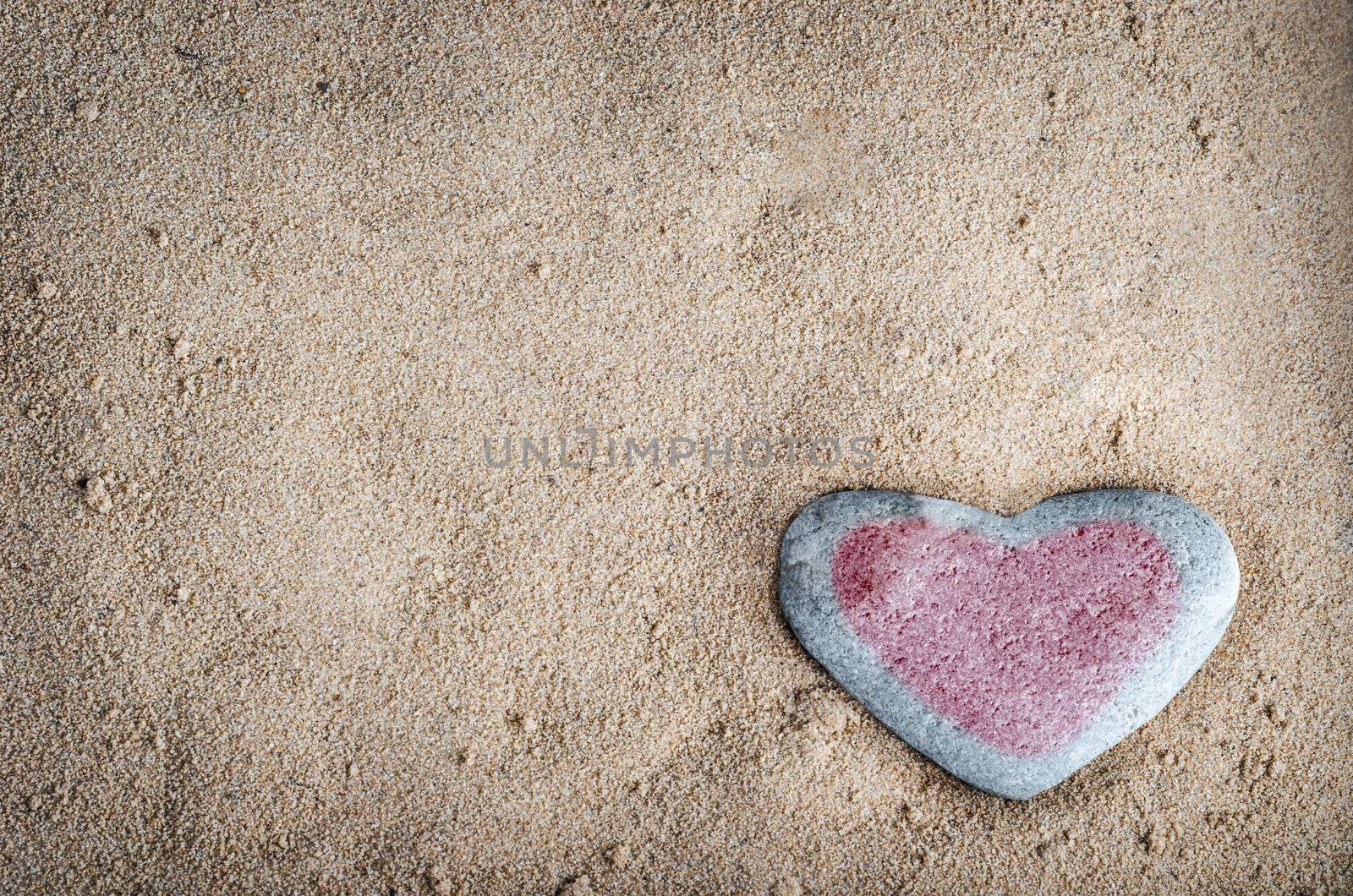 A grey heart shaped stone on grainy sand, tinted with a red heart.  This version is vignetted and edited to give a retro or lo-fi appearance.