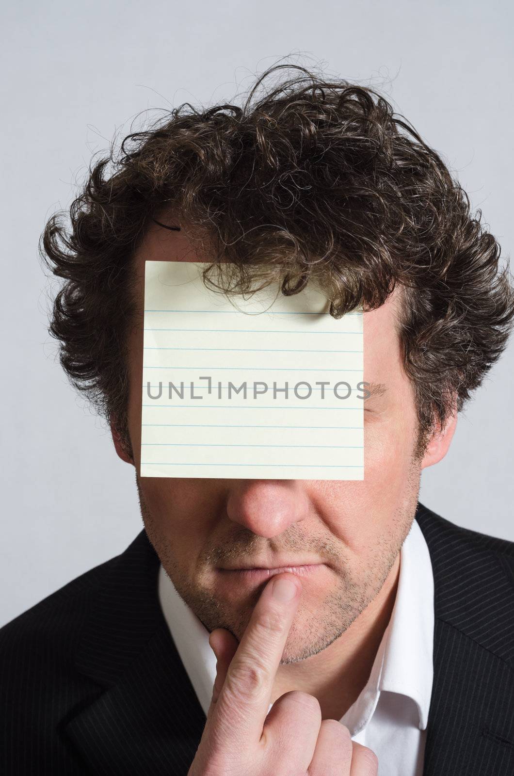 A curly haired man in a suit with shirt slightly open and no tie, resting finger on chin in a contemplative manner, with a blank, lined yellow sticky note on his forehead, facing the camera.