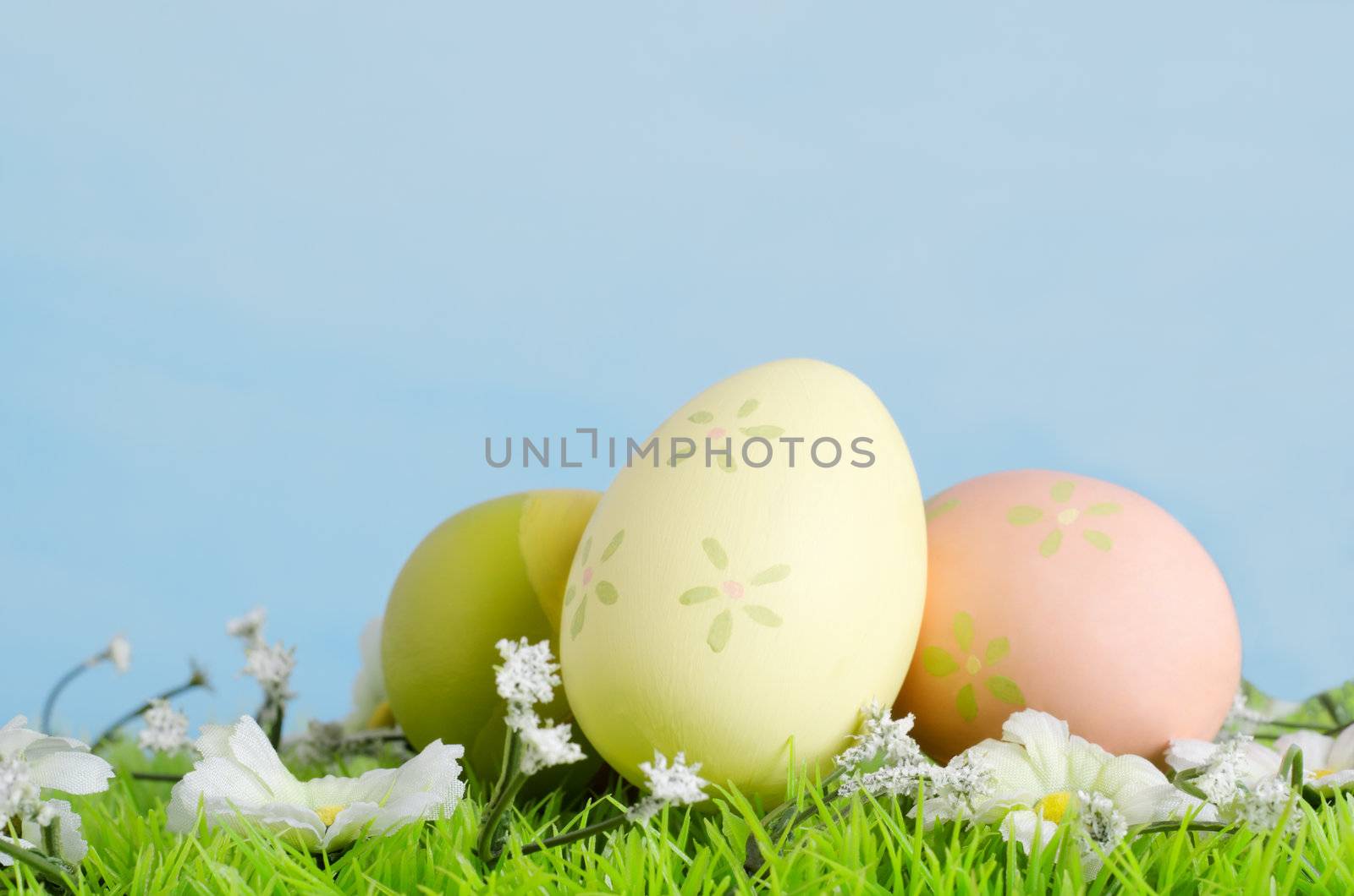 Eggs decoratively painted on artificial grass with fabric flowers photographed against a painted blue background to simulate Spring sky.