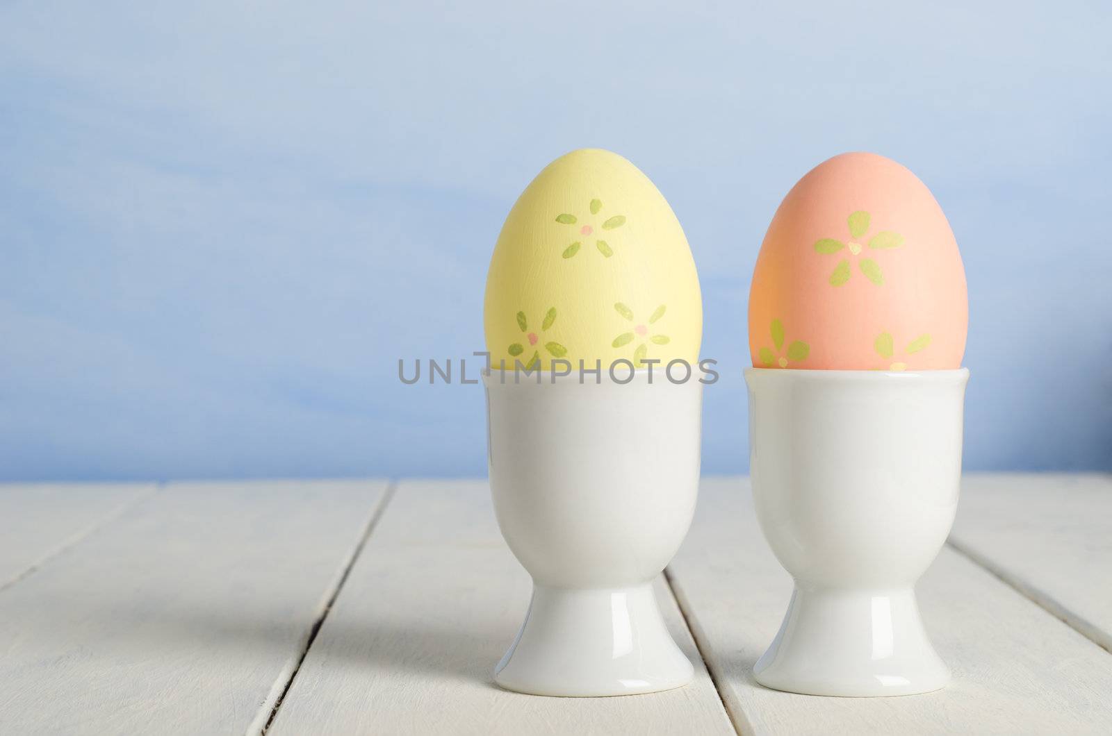 Two handpainted floral Easter eggs  nestled in egg cups on an old cream painted wooden farmhouse table to the right of frame.  Whispy sky blue painted background provides copy space to the left.