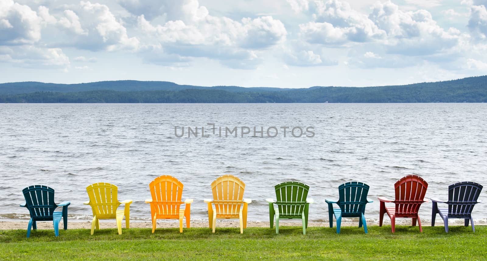 Eight colorful Adirondack chairs lined up on the beach looking out on the lake, mountains and clouds.