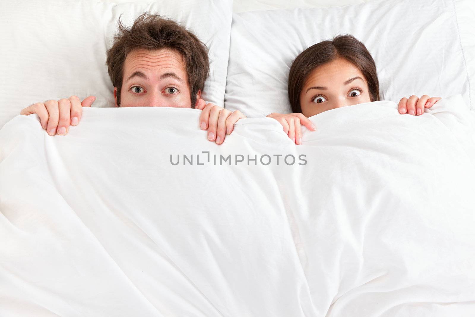 Funny couple in bed looking and peeking over sheets surprised. Young interracial couple, Asian woman, Caucasian man.