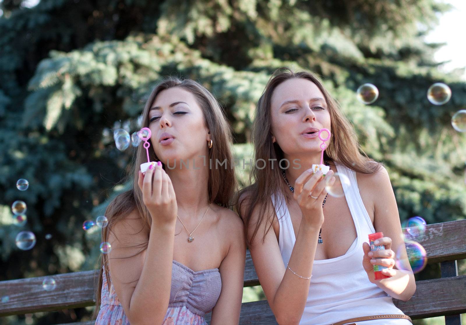 Two beautiful young women make soap bubbles sitting on a bench in park