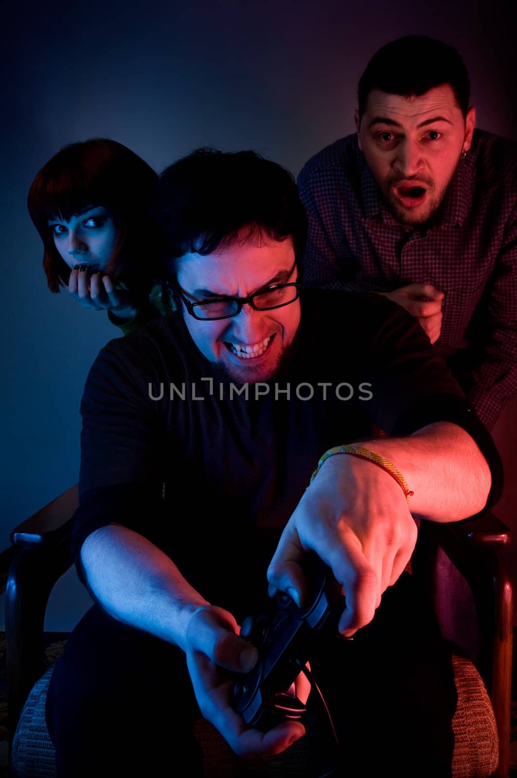 Young emotional man playing video games, two scared persons are hiding behind him 