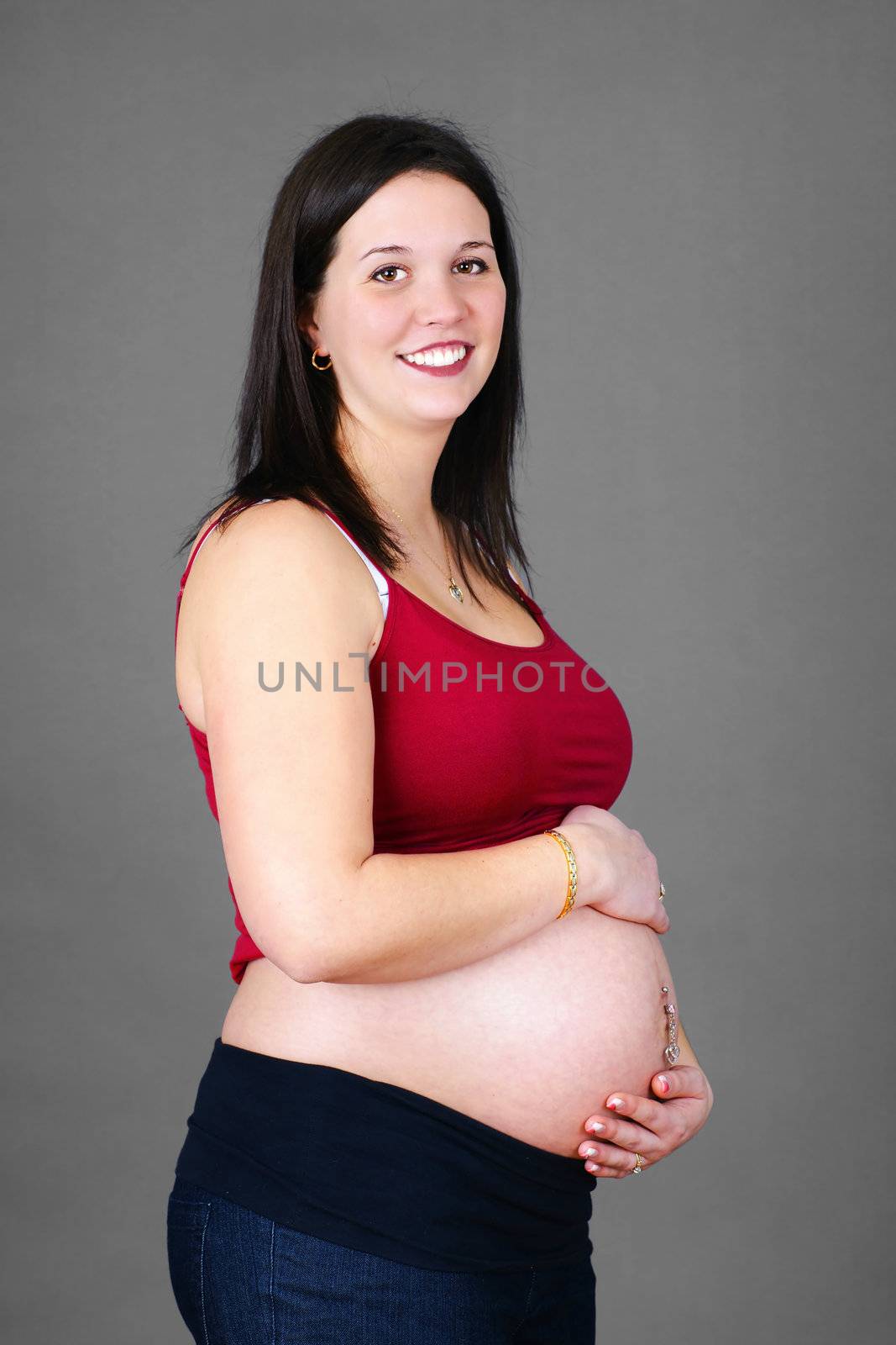 Healthy and naturally beautiful young caucasian mother to be holding her pregnant belly looking at camera, studio shot over grey background.