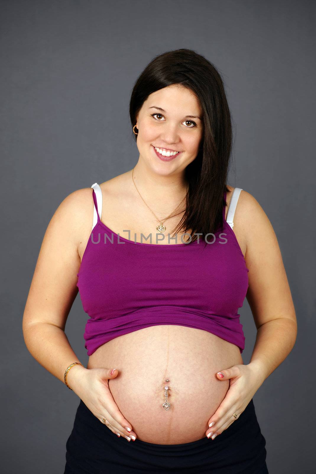 Studio shot portrait of a naturally beautiful, happy and healthy pregnant young woman holding her belly and smiling over grey background.
