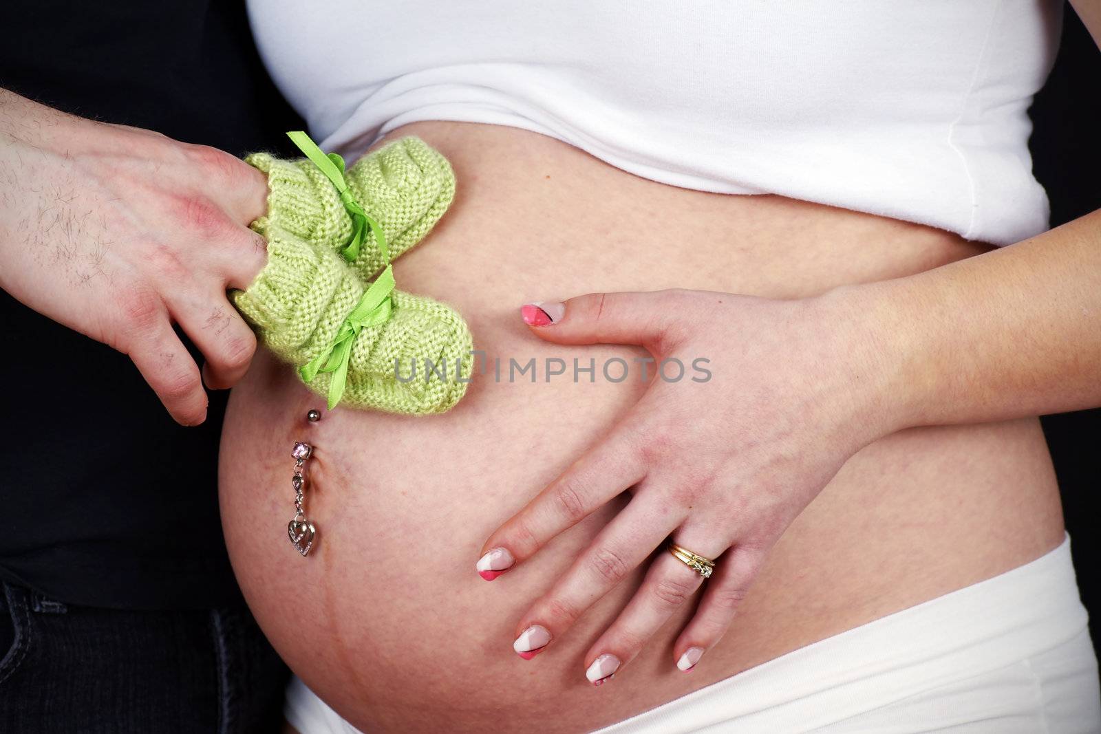 Studio shot of an expecting young married couple with dad to be holding green knit bootees on his wife's pregnant belly.