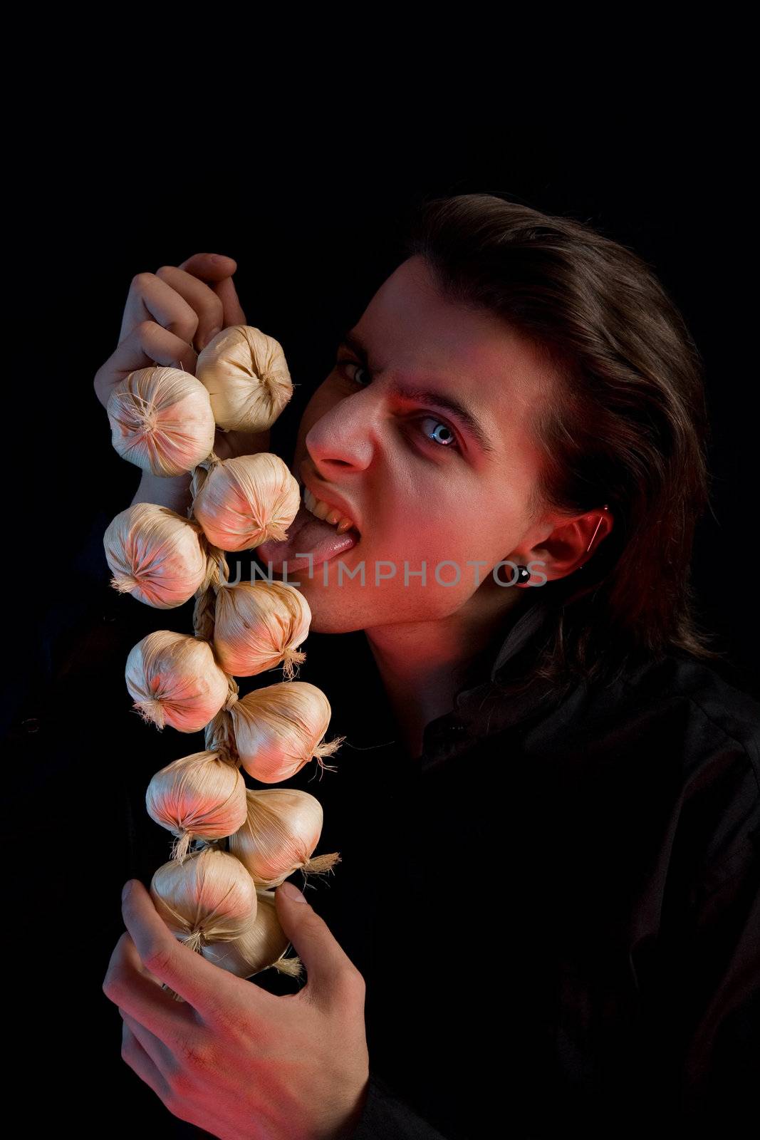 Vampire with scary blue eyes is licking garlic, red horror light, isolated on black background 