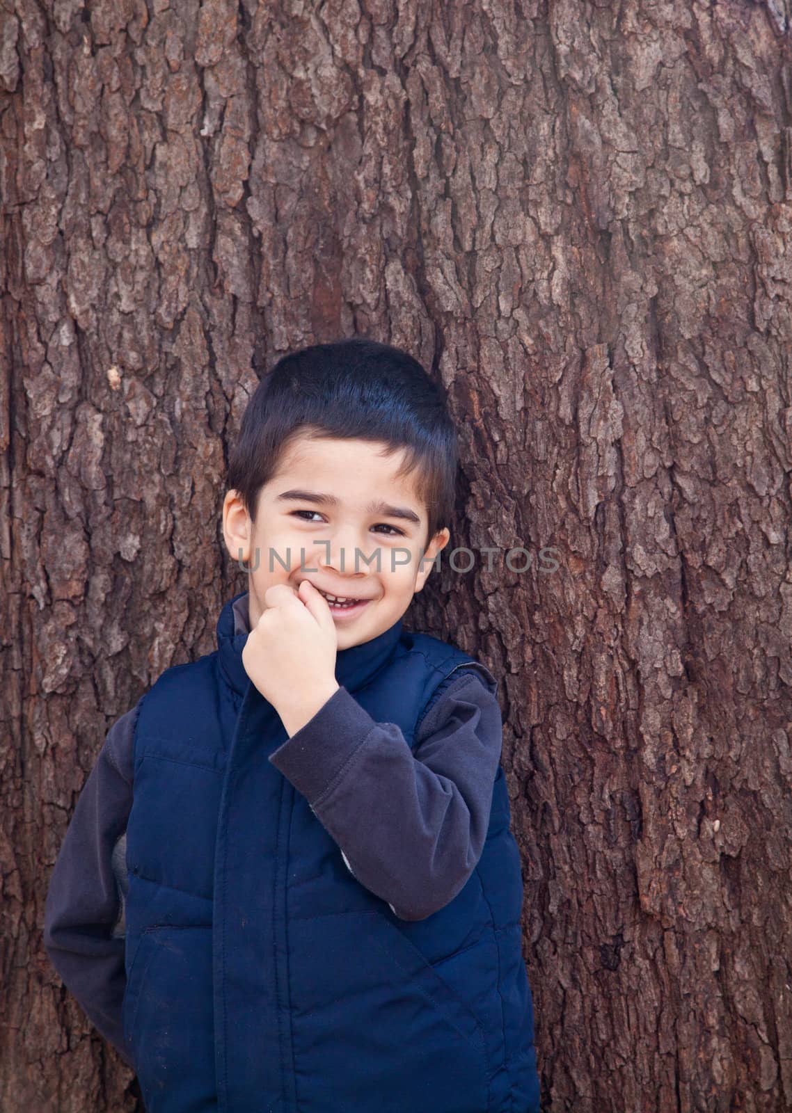 Little boy looking to the left in front of a tree with bark texture in the background biting on his finger with a shy expression.