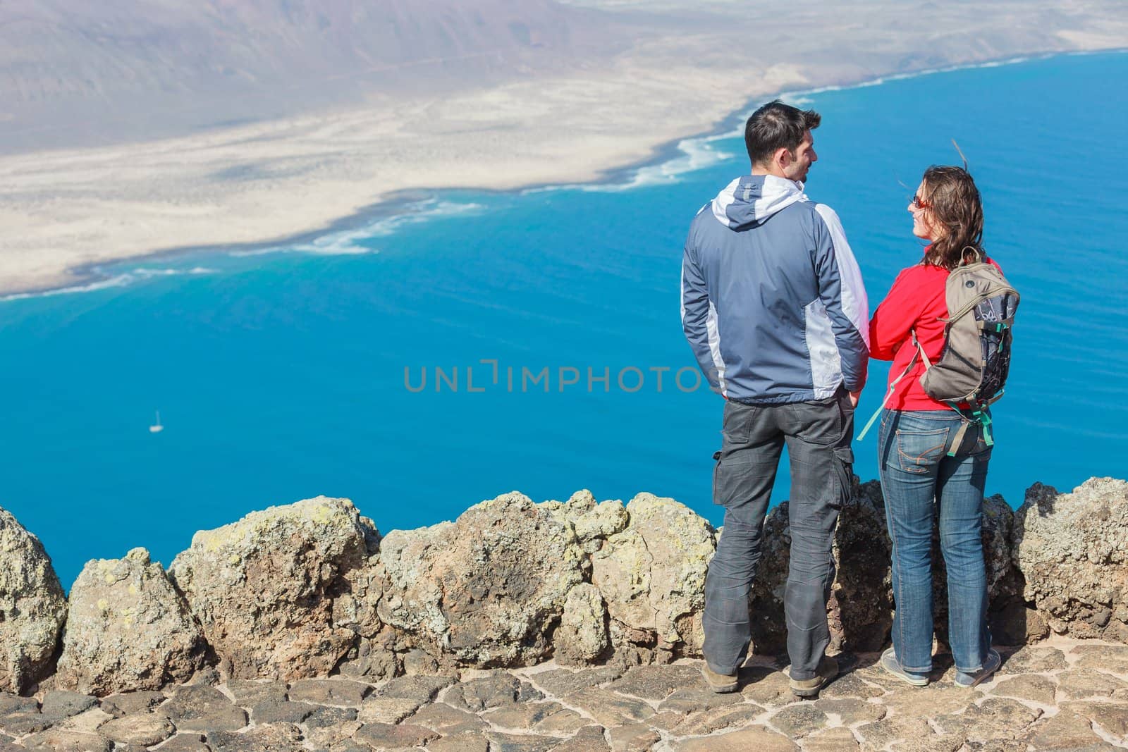 Back view sport couple with backpack standing on cliff's edge and looking to a island.