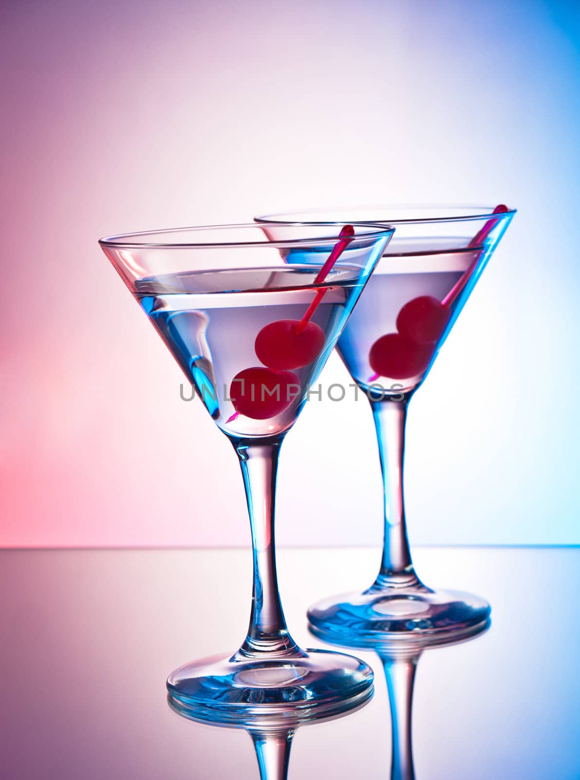 Two glasses of martini with red cherries on a mixed color background