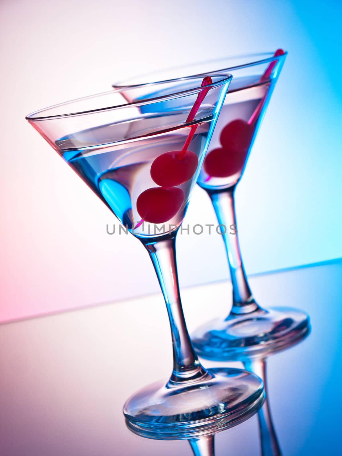 Two glasses of martini with red cherries on a mixed color background