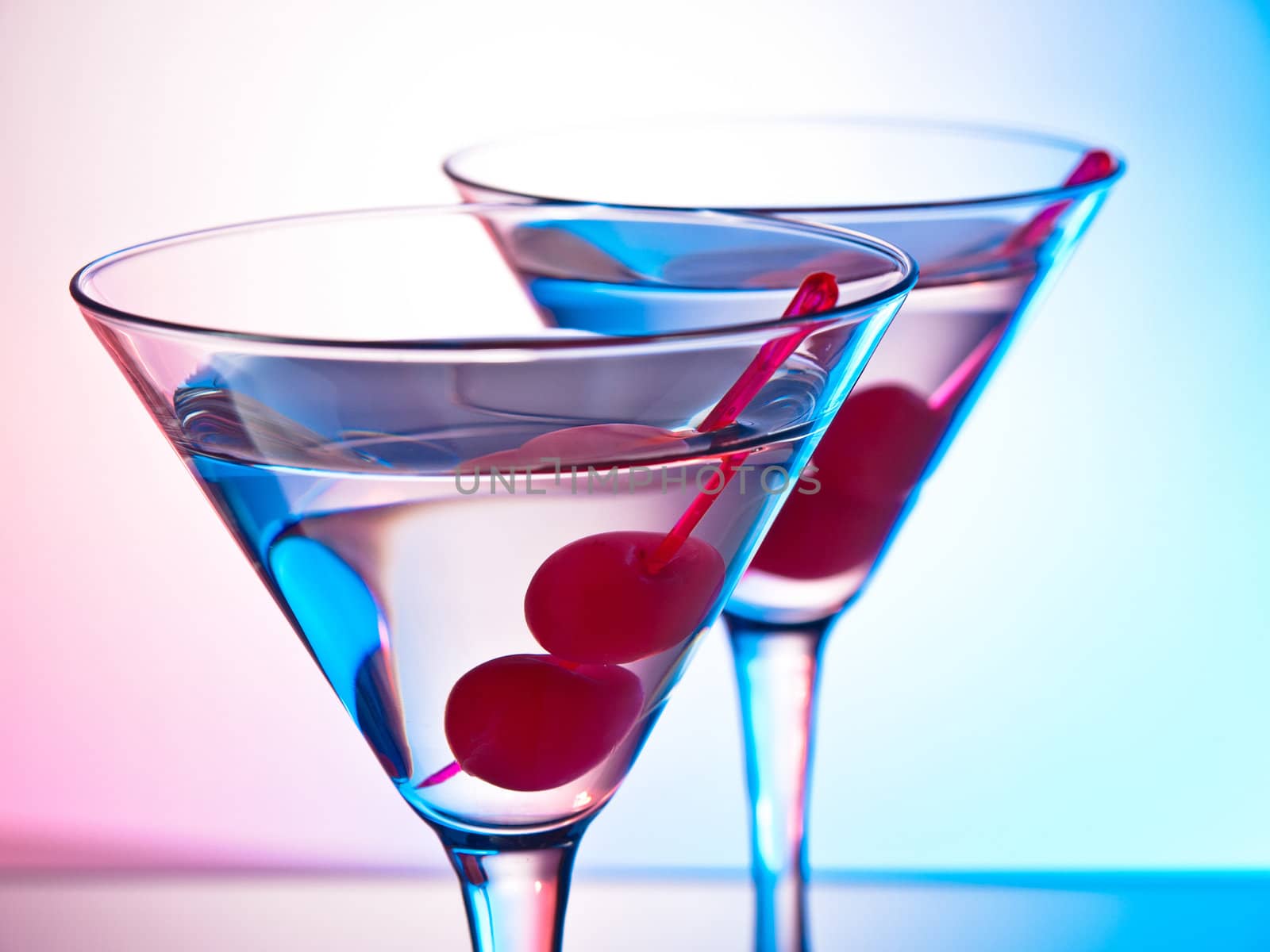 Two glasses of martini with red cherries on a mixed color background