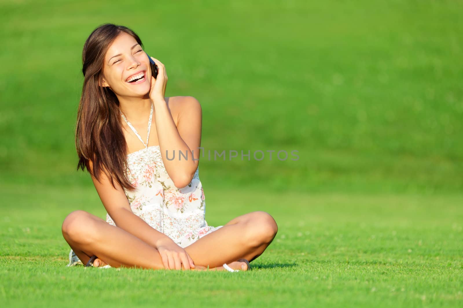 Phone woman laughing in talking in smart phone in park during spring / summer. Happy smiling beautiful young woman sitting in grass. Multiracial Caucasian / Chinese Asian female model outside in summer dress.