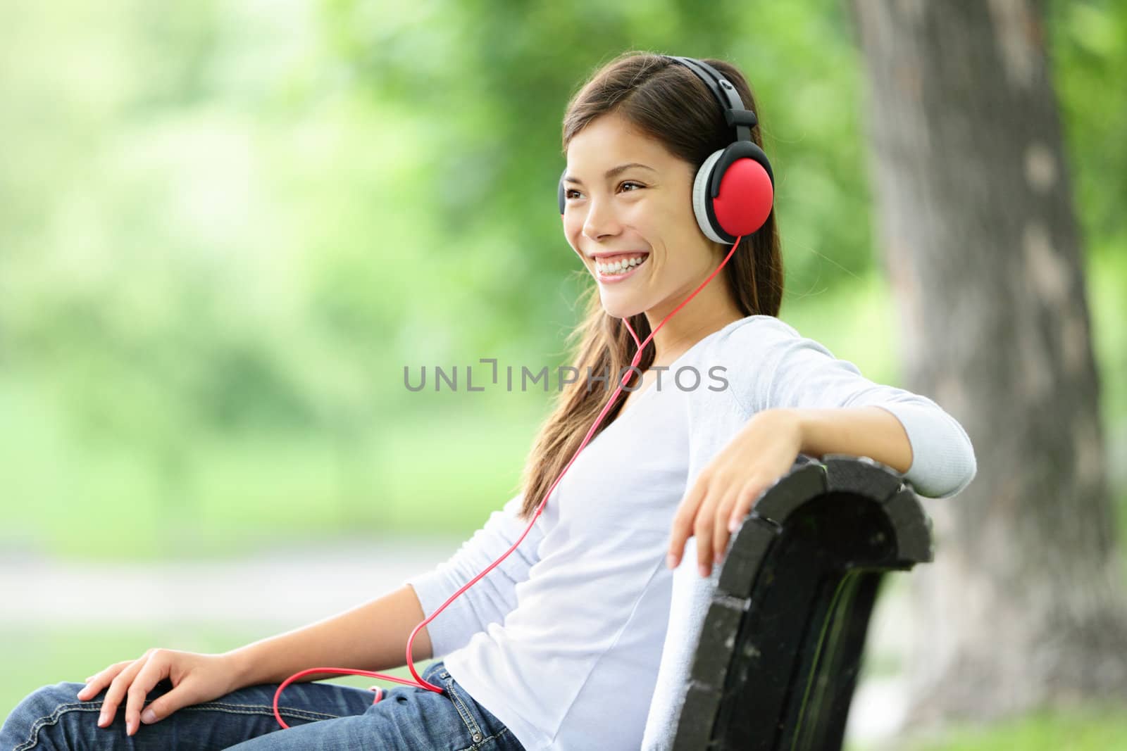 Woman listening to music in park wearing headphones smiling happy. Mixed race Asian Caucasian young woman relaxing outdoors enjoying spring.