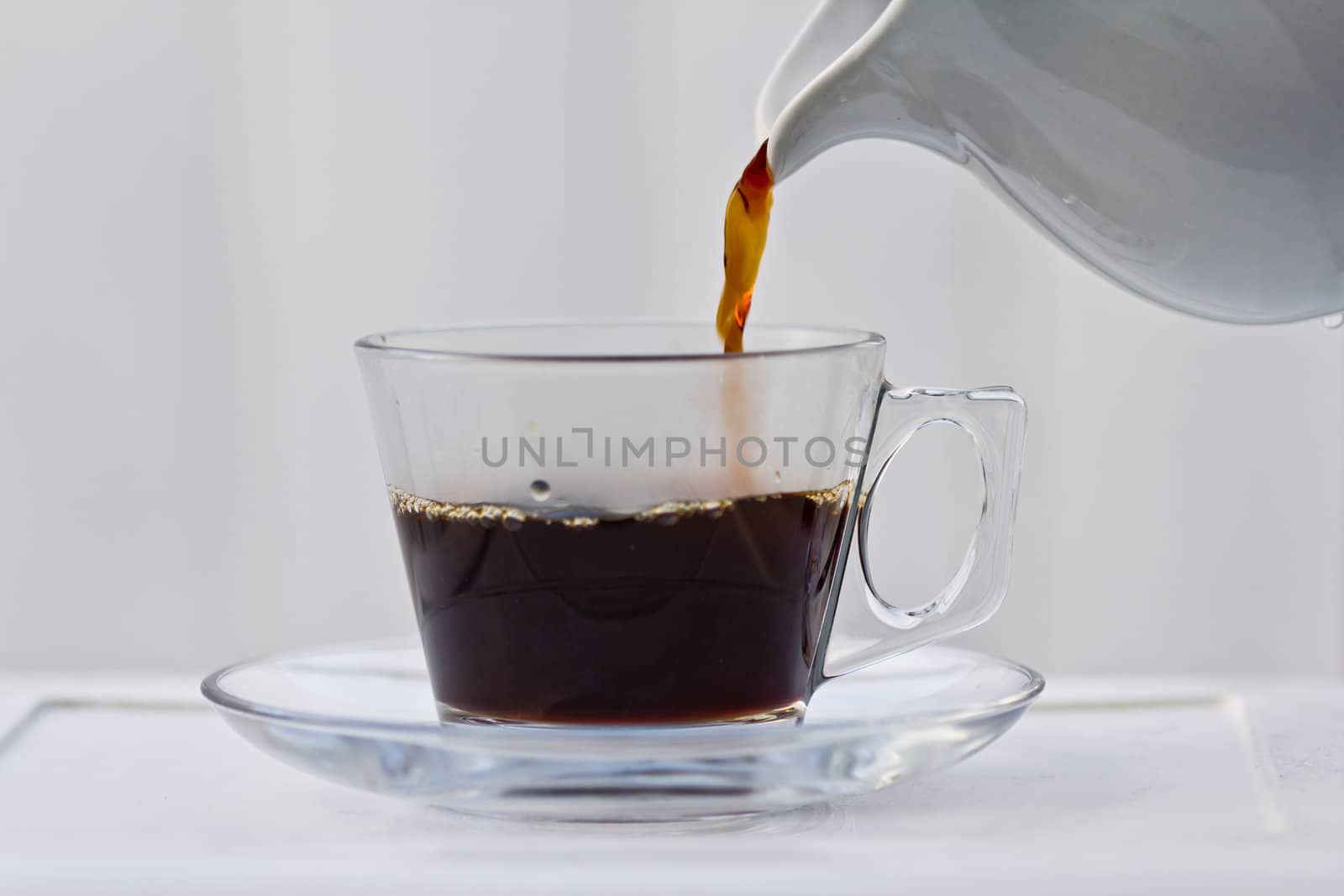 Pouring coffee into a half full clear cup with a saucer.