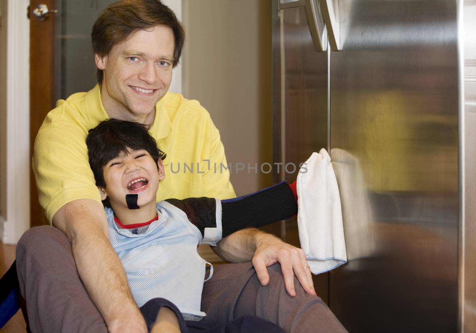 Father on kitchen floor with disabled son, cleaning the fridge. Son has cerebral palsy, with leg orthotics and arm splints.