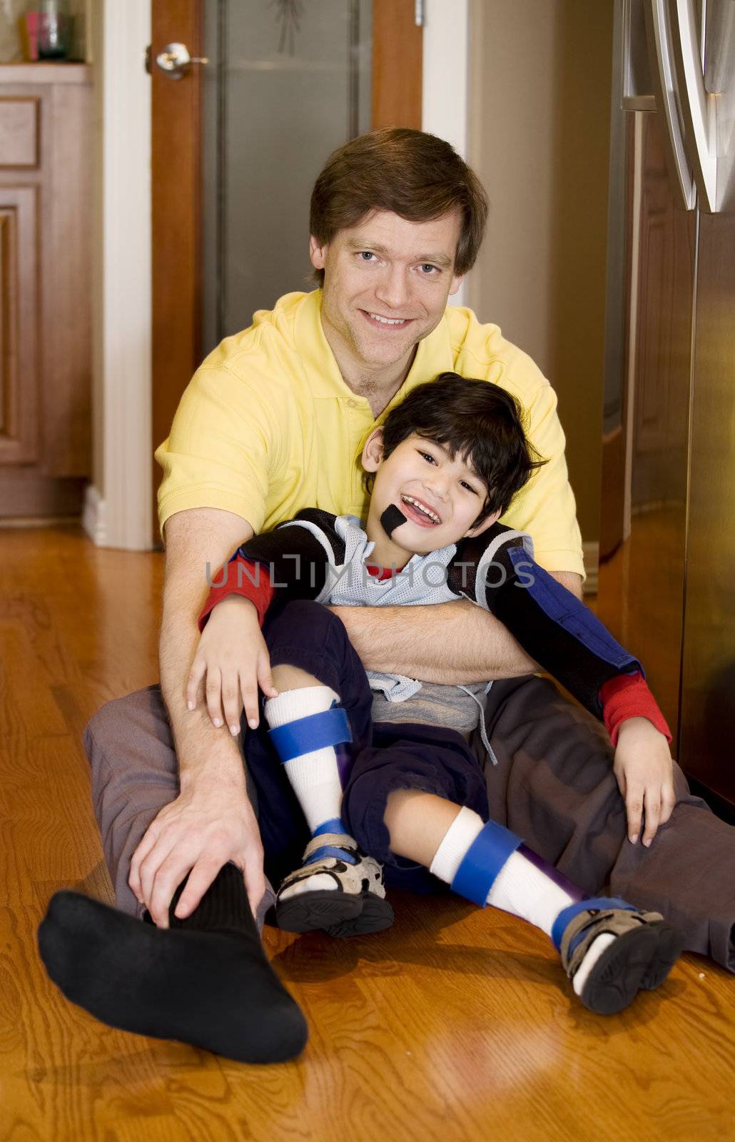 Father holding disabled son on kitchen floor. Son has cerebral palsy.