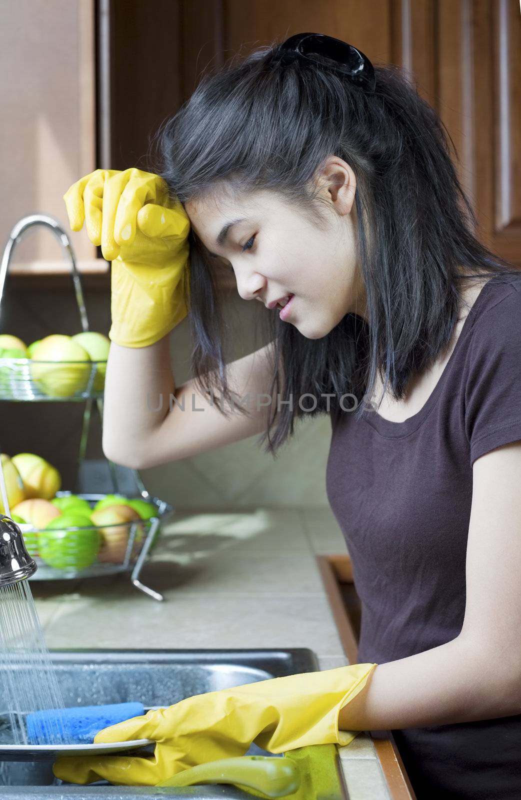 Teen girl washing dishes at kitchen sink, tired expression.