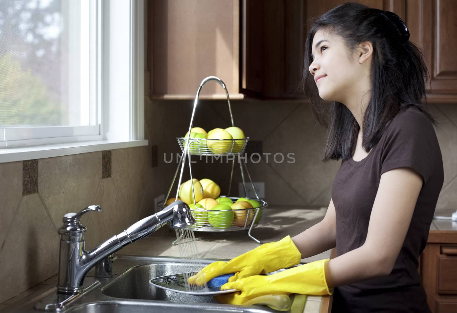 Teen girl washing dishes at kitchen sink, daydreaming or looking out the window with thoughtful expression.