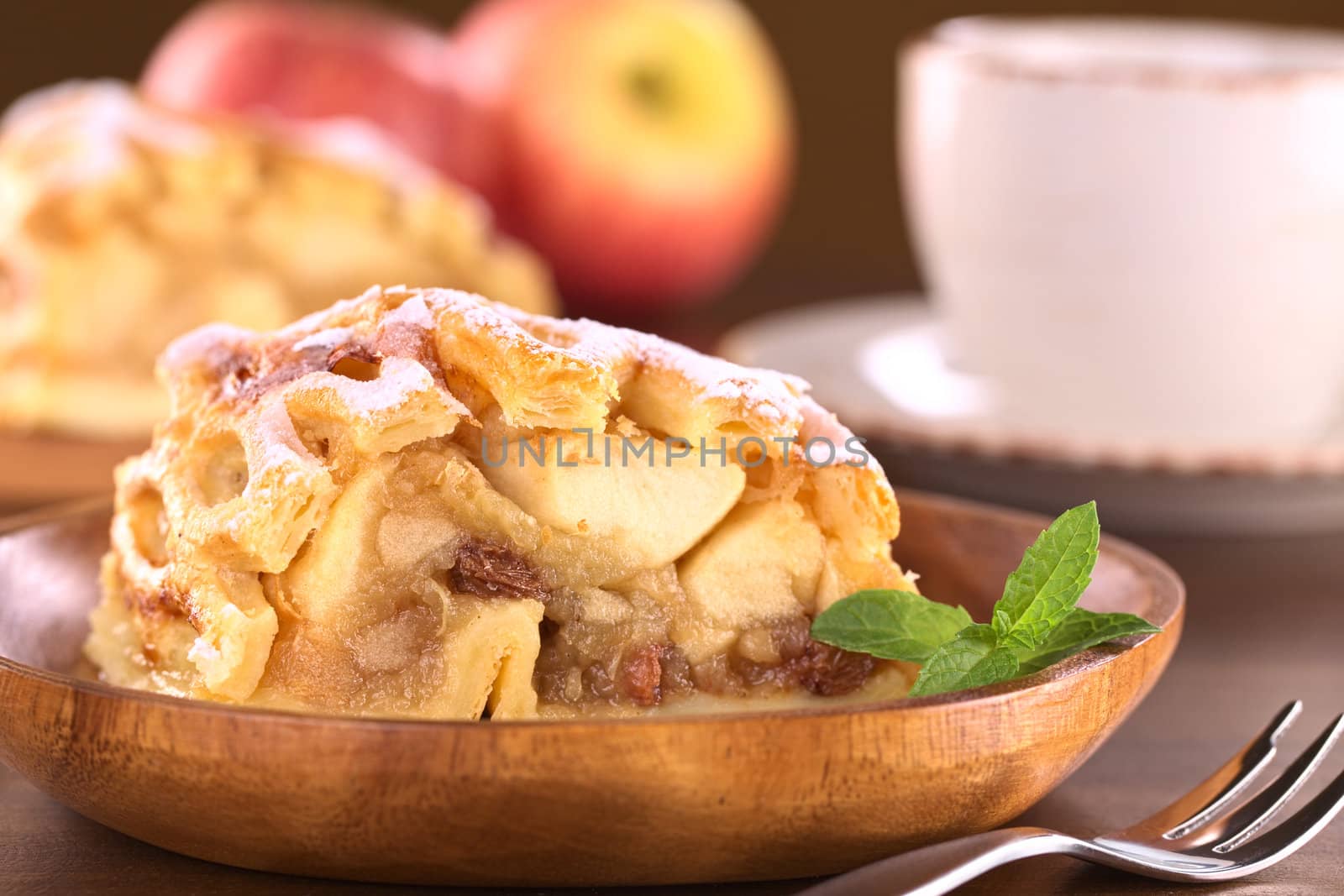 Apple strudel with raisins with coffee cup in back (Selective Focus, Focus on the left side of the apple-raisin stuffing and the front of the mint leaf)