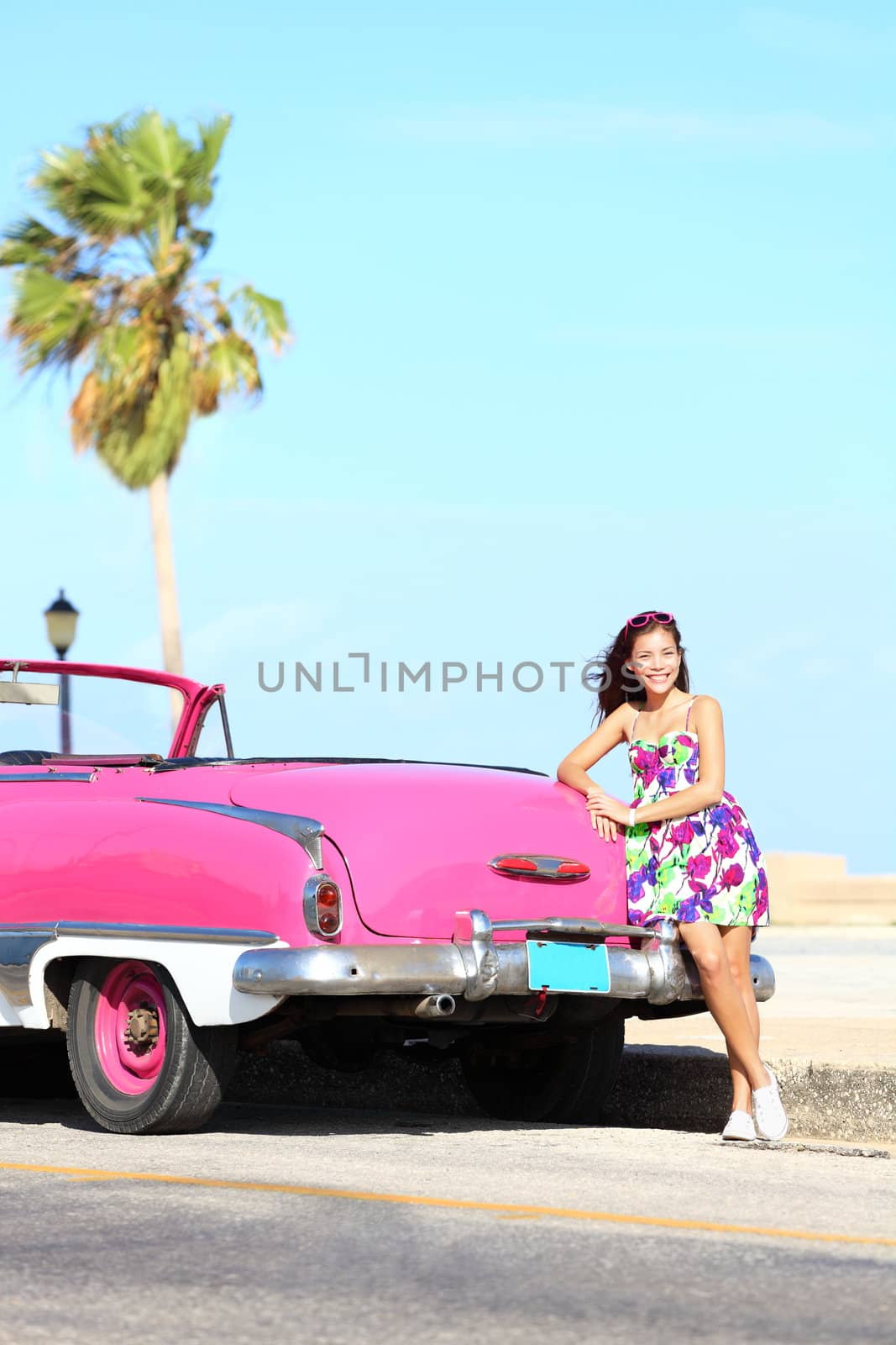 Vintage car and woman standing happy and smiling leaning on pink retro car on the side of the road. Beautiful young multicultural young woman on spring or summer road trip. Havana, Cuba.