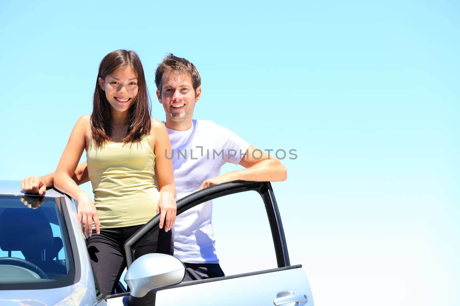 Couple and new car smiling happy outside on sunny summer day during road trip travel vacation. Young interracial couple in their twenties, Asian woman, Caucasian man.