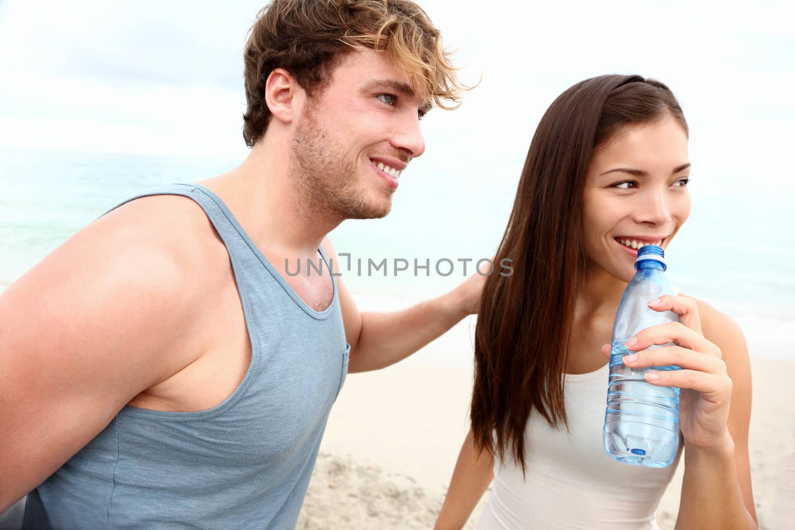 Young couple beach exercise workout. Runners resting taking a break drinking water after running on beach. Young interracial couple, Asian woman fitness sport model and Caucasian man.