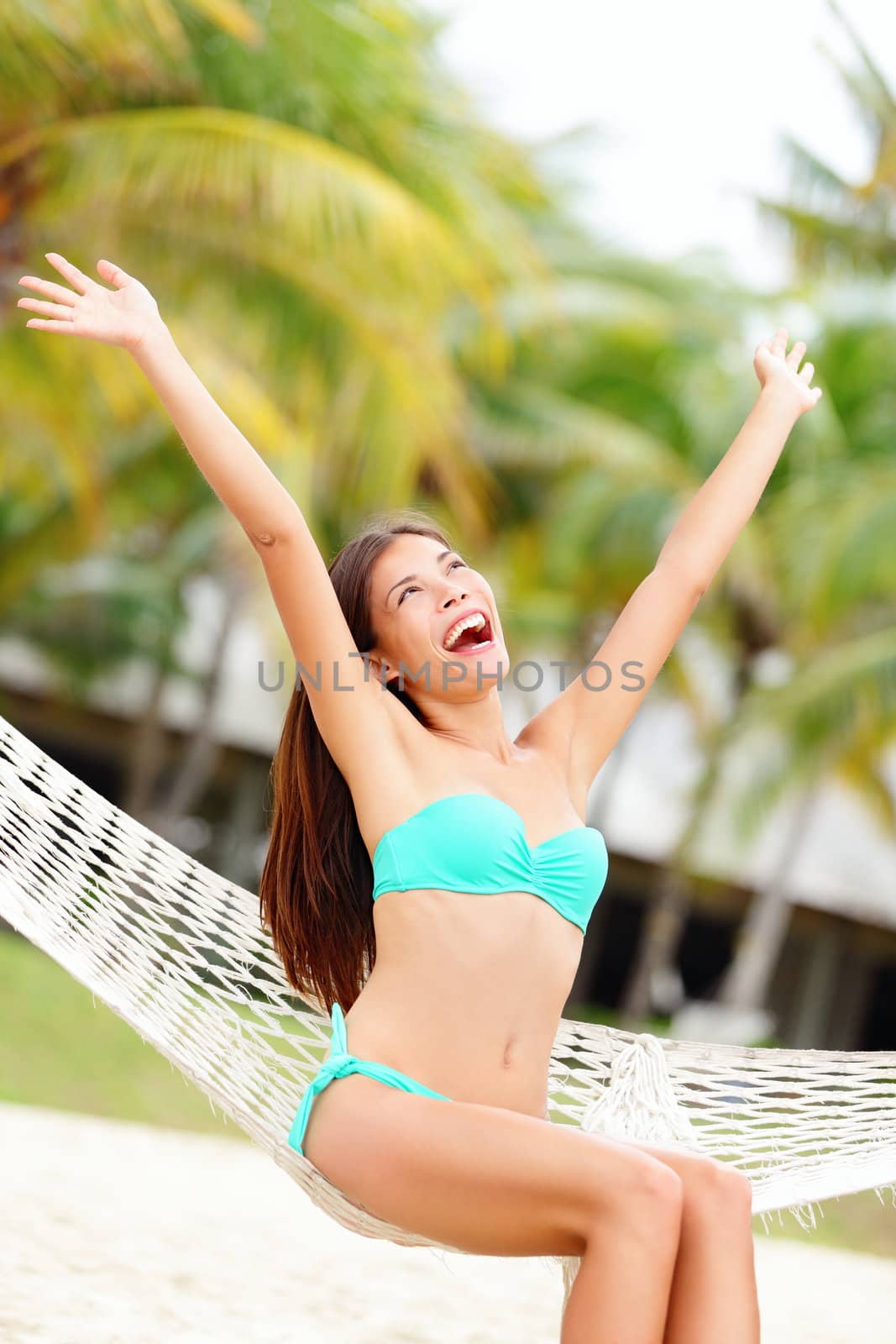 Vacation woman on beach happy and joyful with arms raised sitting in hammock. Beautiful young bikini model in summer holidays resort. Mixed race Caucasian / Chinese Asian woman.