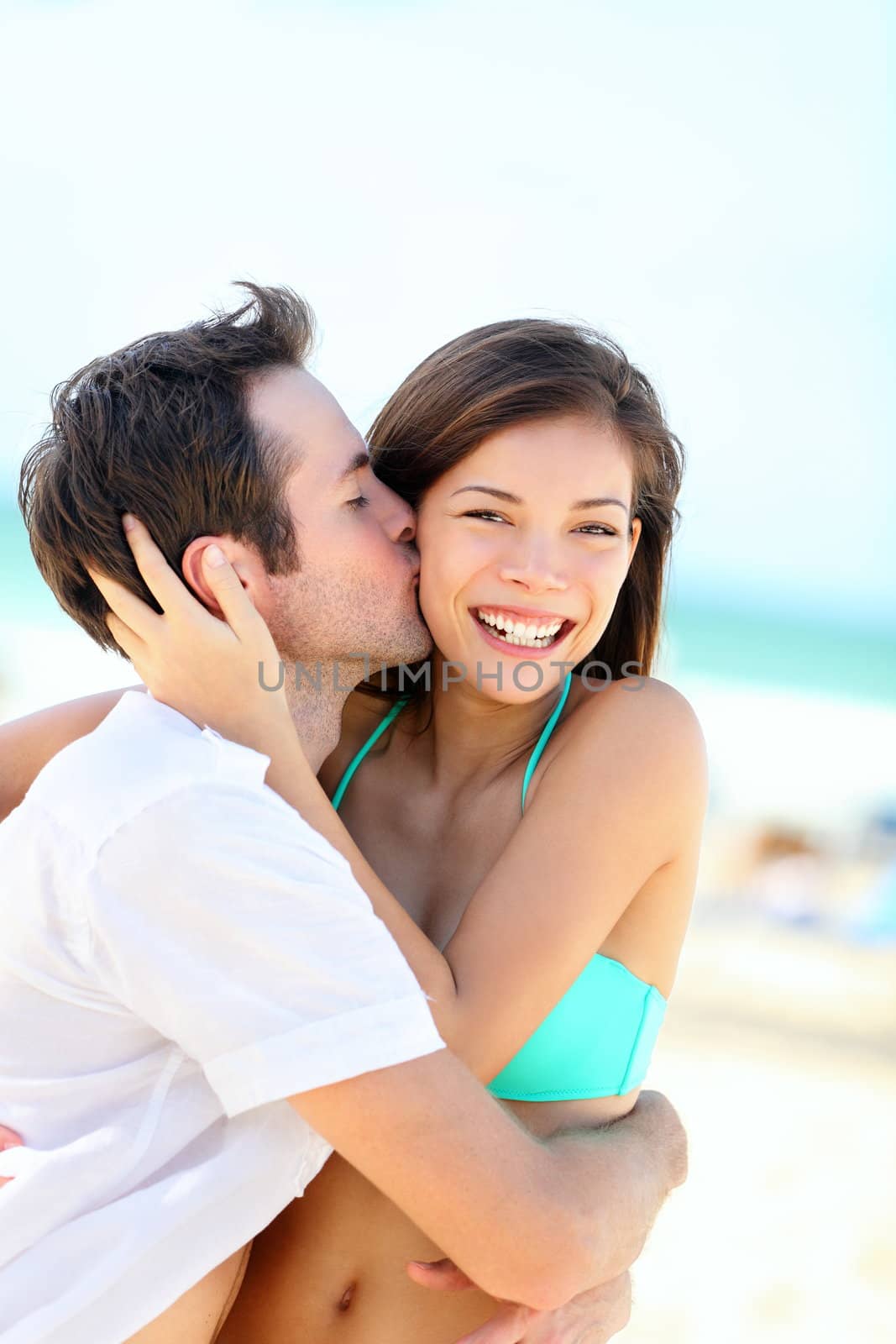 Happy couple kissing and embracing in joyful happiness showing love during summer beach vacation. Beautiful young interracial couple, Asian woman, Caucasian man outdoors.
