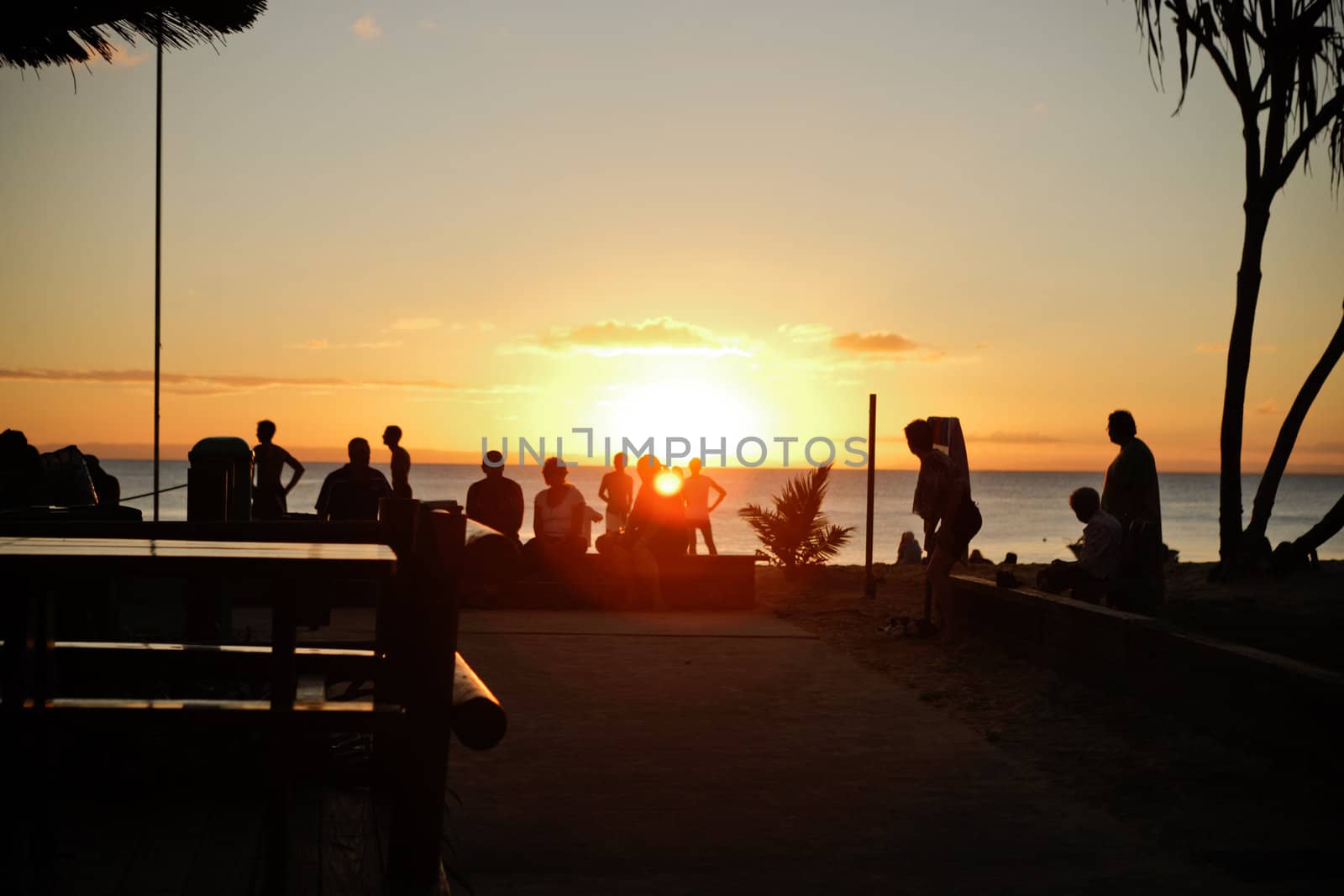 People on a tropical beach at sunset silhouetted against the orange glow of the setting sun as it drops below the horizon of the ocean