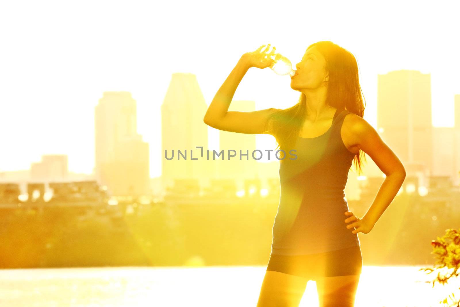 Summer runner woman drinking water in sunshine on warm sunny day in city park with cityscape skyline in background. Beautiful young female fitness runner relaxing after outdoor workout in Montreal, Quebec, Canada. Multiracial woman jogger.