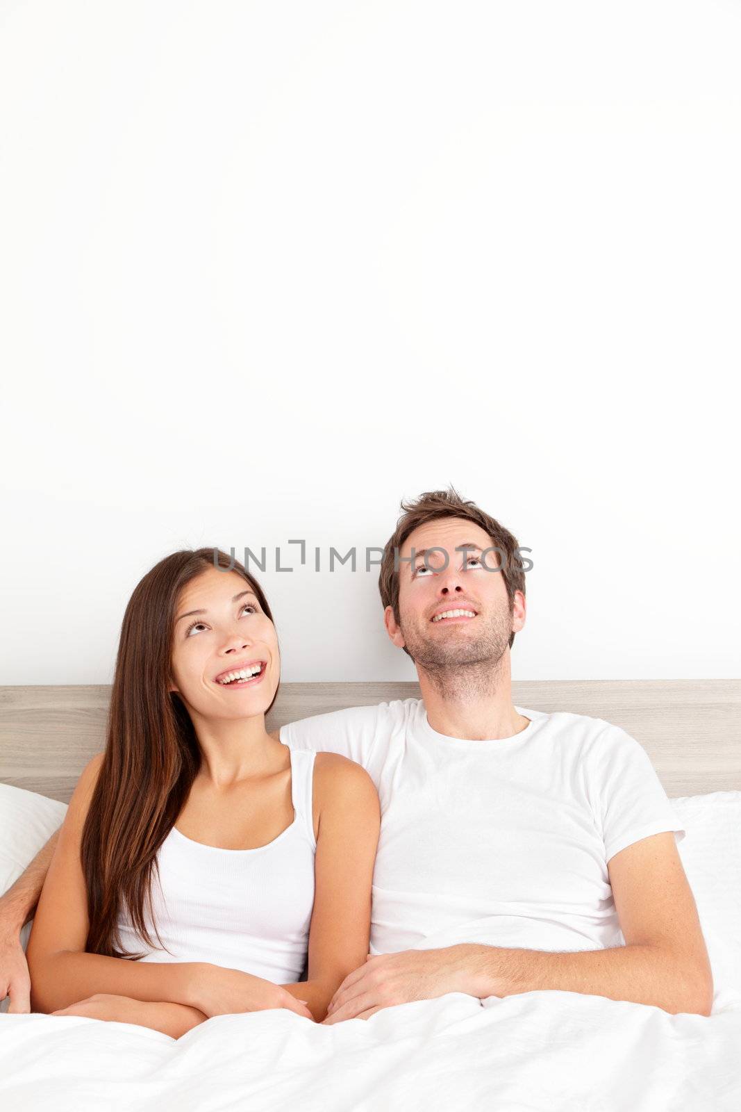 Couple thinking in bed looking up showing copy space. Happy young couple together in bed day dreaming of future. Caucasian man, Asian woman.